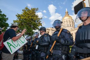 A row of police officers in riot gear is confronted by a protester holding a sign that say "ACAB"