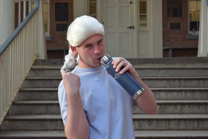 Photo of Lawrence Trevette twisting a white founding fathers wig and sipping from a blue-gray Alexander Hamilton travel mug
