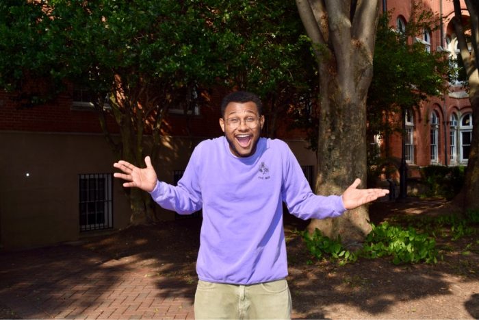 Photo of Will Hammond wearing a purple shirt and smiling while enthusiastically shrugging in Dahlgren Quad