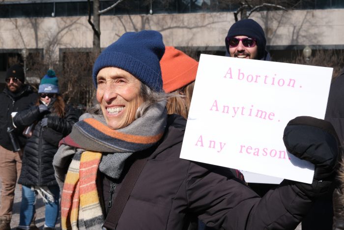 An elderly woman wearing a blue hat and colorful scarf is smiling and holding a sign that is white and in pink text reads "Abortion, anytime, any reason."