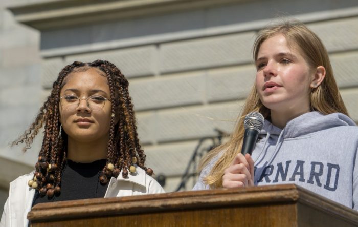 Elizabeth Foster speaks into a microphone at a podium in front of a white brick background. Another student activist stands beside her.