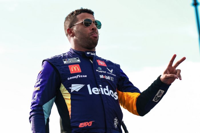 An image of Bubba Wallace from a lower angle, as he flashes a peace sign to an audience off screen.