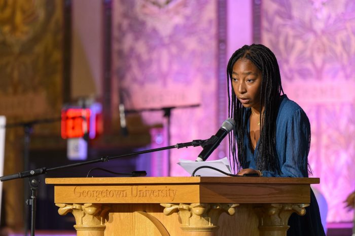 Kessley Janvier is shown speaking into a microphone at a podium labeled "Georgetown University" in Gaston Hall. The background is tinted pink.