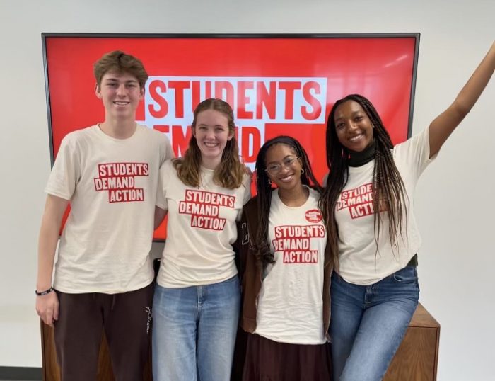 Emma Vonder Haar is pictured with three other students, all in white t-shirts with "Students Demand Action" written on them in red lettering. The students are standing in front of a TV screen with the Students Demand Action logo on a red background.