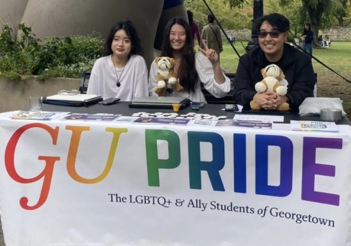 Gisell Campos sits on with two other students on their right, at a table with a white banner that reads "GU Pride" in rainbow lettering. Campos and the student in the middle are holding Jack the bulldog stuffed animals.