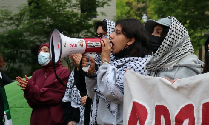 Selina Al-Shihabi speaks into a megaphone at a protest. She is wearing a keffiyeh and standing with other organizers and demonstrators.