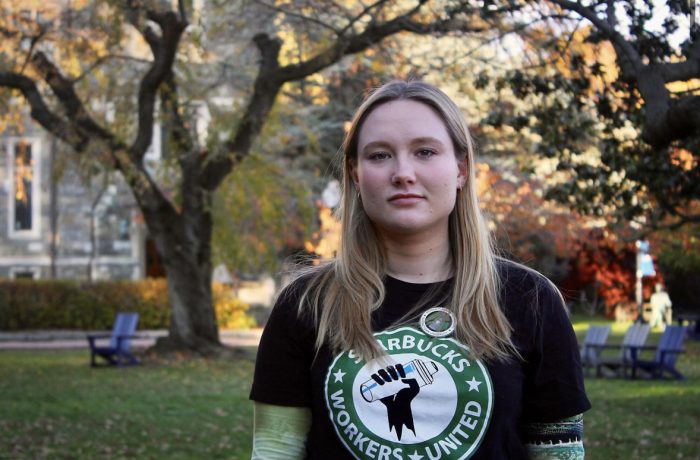 Elinor Clark looks into the camera in a headshot photo taken on Copley Lawn. Clark is pictured in front of a tree and White-Gravenor Hall. She is wearing a black Starbucks Workers United t-shirt.