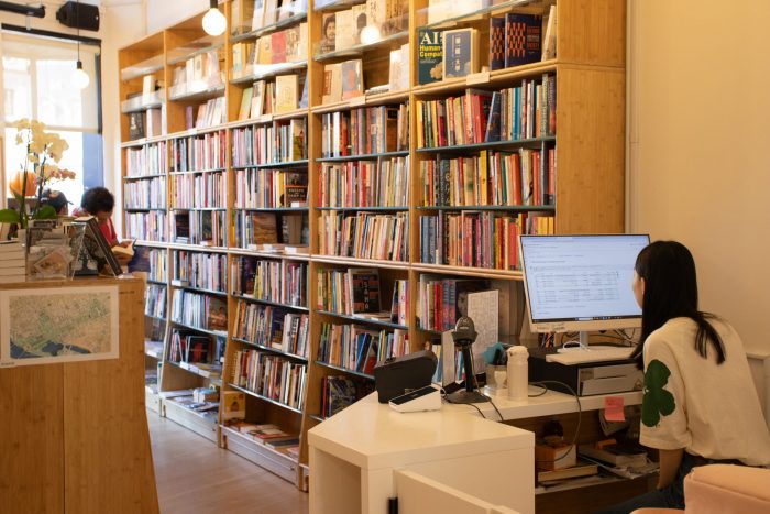 A cashier sits at a computer. The cashier is facing a bookshelf along the wall.