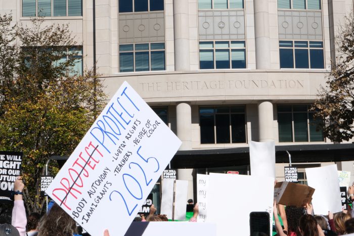 Protesters stand in front of the Heritage Foundation. Next to the entrance, a sign reads, “Project (strikethrough) Protect bodily autonomy, LGBTQ rights, immigrants, teachers, people of color 2024”