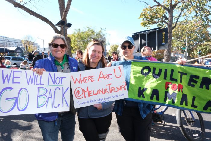 Gosse and Harris smile and pose alongside another woman while holding signs that say, “We will not go back,” “Where are U @MelaniaTrump?,” and “Quilters for Kamala”