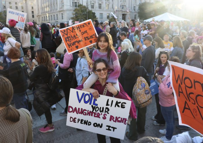 A young girl sits on her mother's shoulders. The girl is carrying a handwritten sign that reads, "Women's rights are human rights." The mother's sign reads "Vote like your daughter's rights depend on it."