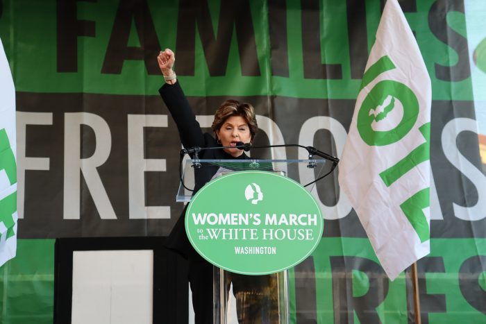 Gloria Allred speaks at a podium with a green "Women's March to the White House" sign on it.