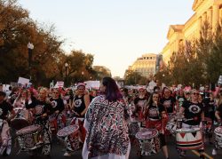 Bátala Washington D.C., an all-female Afro-Brazilian percussion band, marches in the Women's March. They are playing drums and wearing red, white, and black clothing.