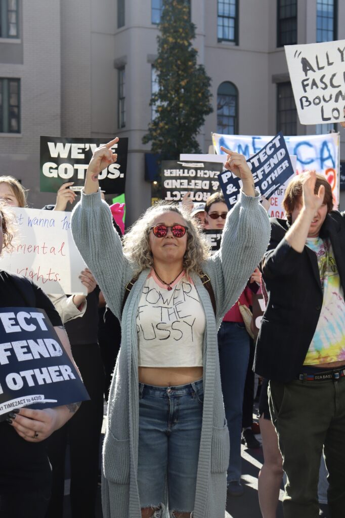 A woman holds both of her middle fingers in the air in the middle of the rally, wearing a shirt that says, “Power of the Pussy”