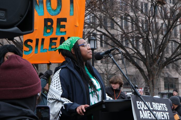 Nachega wears a green bandana on her head and a keffiyeh over her shoulders while speaking at a podium with the words ‘Equal Rights Amendment’ written on a sign in the front