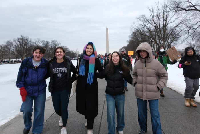 Five girls, all wearing coats and jeans, walk along the sidewalk hand-in-hand with the Washington Monument in the background