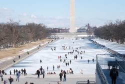 The Washington Monument is pictured in the background, with the frozen reflecting pool in front of it. Visitors walk on the frozen pool and on the sidelines.