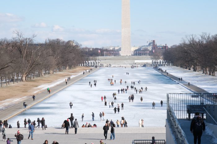 The Washington Monument is pictured in the background, with the frozen reflecting pool in front of it. Visitors walk on the frozen pool and on the sidelines.