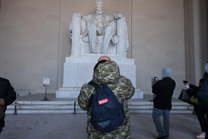 A visitor wearing a camoflauge jacket and a black backpack with MAGA written on it in red and white stands in front of the Lincoln statue in Lincoln Memorial.