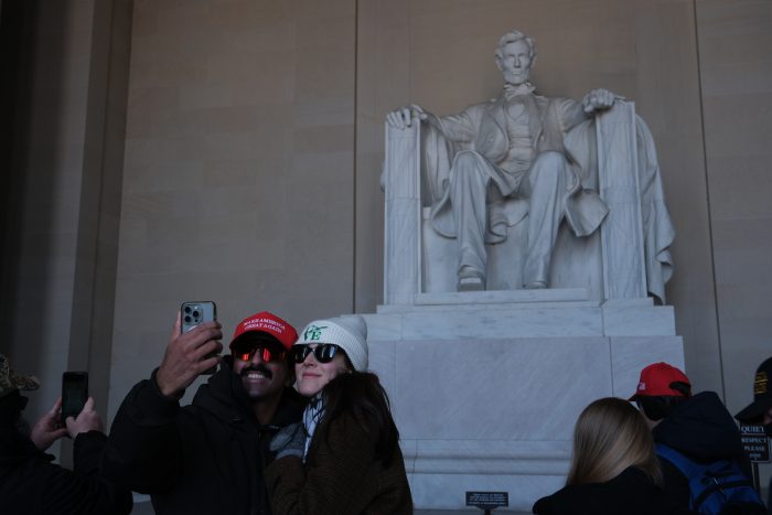 Two people take a selfie in front of the Lincoln statue. One is wearing a white beanie, while the other wears a MAGA baseball cap.