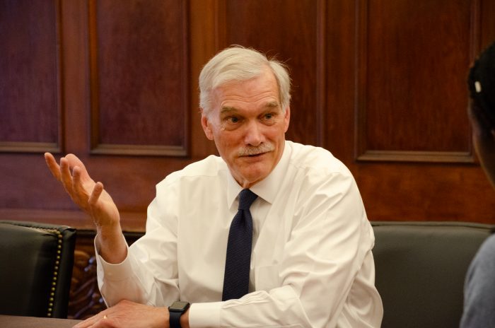 Interim President Groves sits at a table, gesturing with his hand as he speaks. He is wearing a white shirt and navy tie.