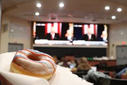 A white donut with red and blue stripes is held up on a napkin. The inauguration plays on two large screens in the background.