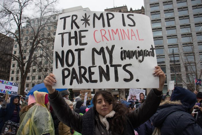 Mejia, wearing a black coat and a white scarf, holds up a sign with the words “Tr*mp is the criminal not my immigrant parents”