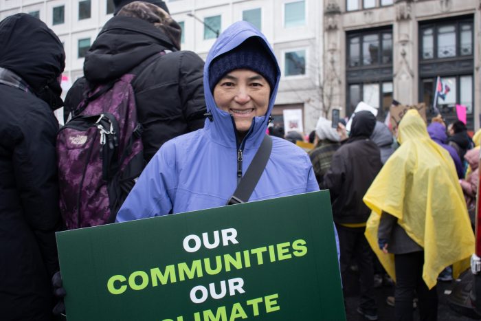 Phuong Hoang poses in a purple jacket with a green sign reading ‘Our Communities, Our Climate’