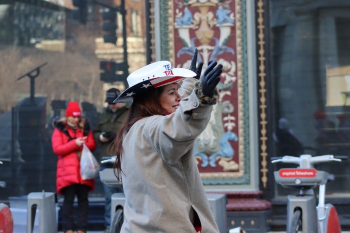 A person wearing a white hat with "Team Trump" written on it raises their arms to the air.