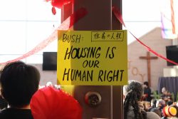 A handwritten poster that reads "BUSH: HOUSING IS OUR HUMAN RIGHT" on yellow paper hangs on a post inside. Red streamers and lanterns are also hanging off of the post.