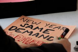 Someone's arm is pictured writing on a poster that reads "NEW YEAR SAME DEMANDS NO DISPLACEMENT" on orange paper