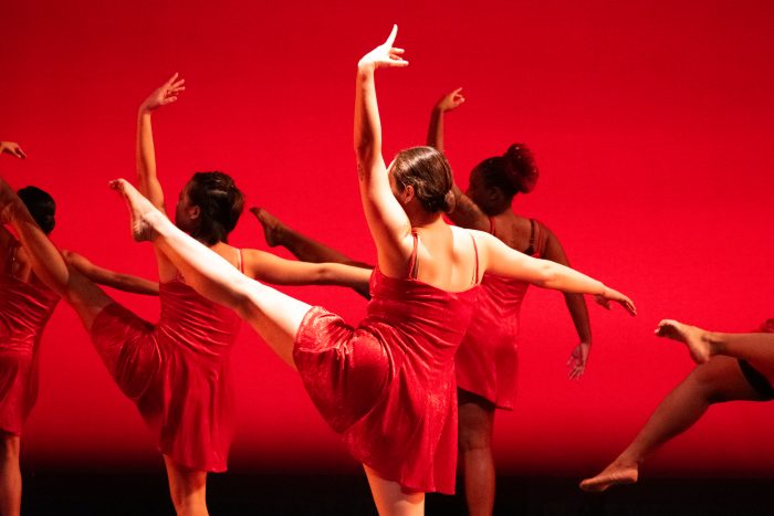 Dancers with red dresses face backwards, with their left legs extended in the air.