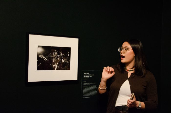 A person in a dimly-lit room speaks while looking at an image mounted on the wall. The image shows people with instruments.