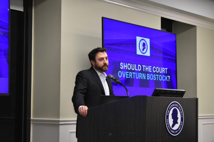 Josh Hammer speaks at a podium that reads, "The Federalist Society: Debate, Discuss, Decide." Behind him is a television screen with the title of the debate, "Should the Court Overturn Bostock?"