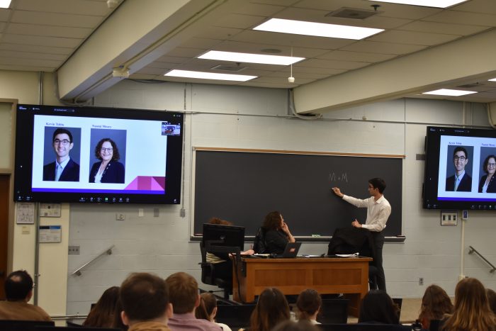 Kevin Tobia writes on a chalkboard while Naomi Mezey sits next to him at a desk in the front of a classroom. TVs next to him have pictures of Tobia and Mezey on them. Students sitting in rows listen to Tobia's remarks.