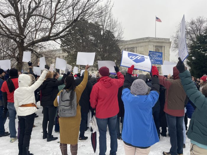 A crowd of people holding signs, facing away from the camera, rallies outside. It is an overcast day.