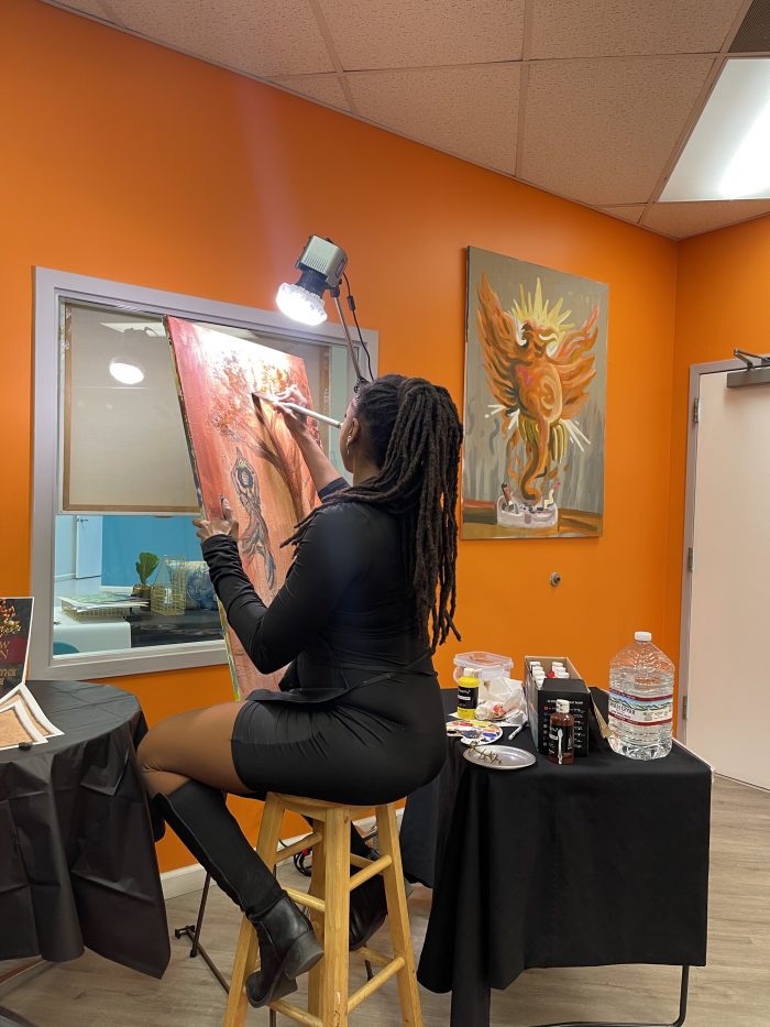 An artist, facing away from the camera, paints on an easel. The room has orange walls. The painting has an orange background and shows a figure and a tree.