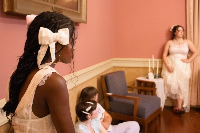 A Black girl (Fife) with long braided hair and bows sits watching a model pose