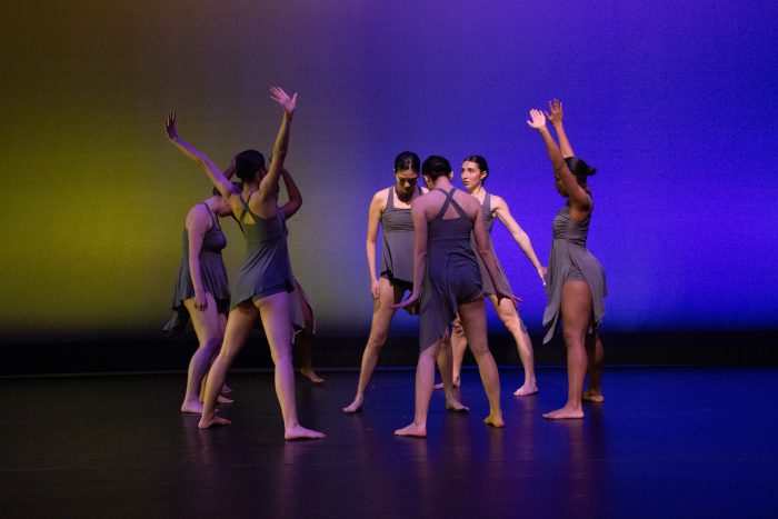 A circle of seven dancers in grey outfits, with varying arm poses.