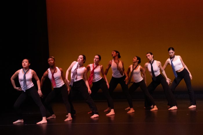 A diagonal line of eight dancers, in white tops, black pants, and varying ties, pose together.