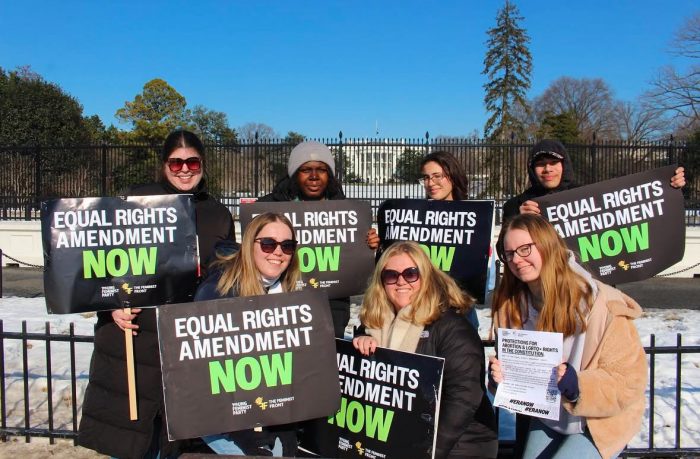 A group of seven people stand or kneel outside the black gates around the White House. They are holding signs that read, "Equal Rights Amendment Now."