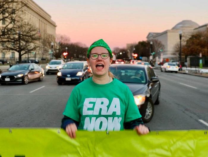 A person wearing a green T-shirt that reads "ERA Now" stands in the middle of a street with cars, stopping traffic. The person appears to be yelling, and is holding a large neon green banner.