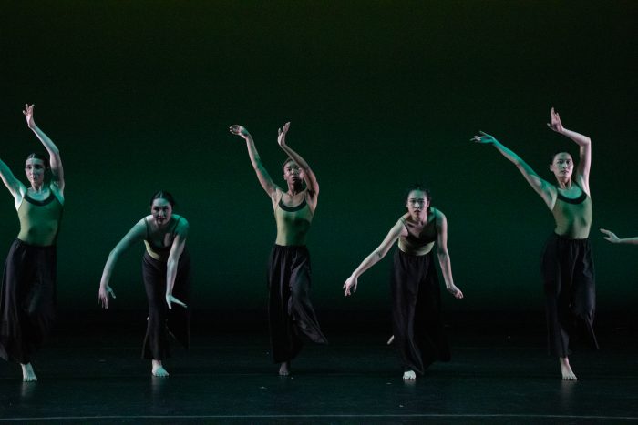 A line of five dancers in green tops and black pants alternate in arm poses.