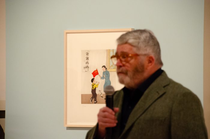 A person speaks into a microphone. In the background, there is a painting of an adult handing a child a Chinese flag.