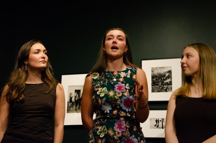 Three people facing the camera stand in a dimly lit room in front of three images mounted on the wall. The person in the middle is speaking.