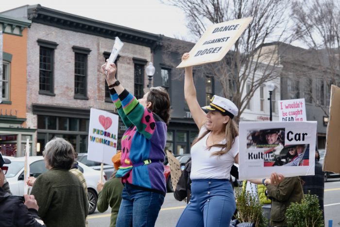 Steffens, wearing a multicolored sweater and jeans, and Knutson, wearing jeans, a white shirt, and a boat captain’s hat, dance on top of a bench while holding signs up in the air.