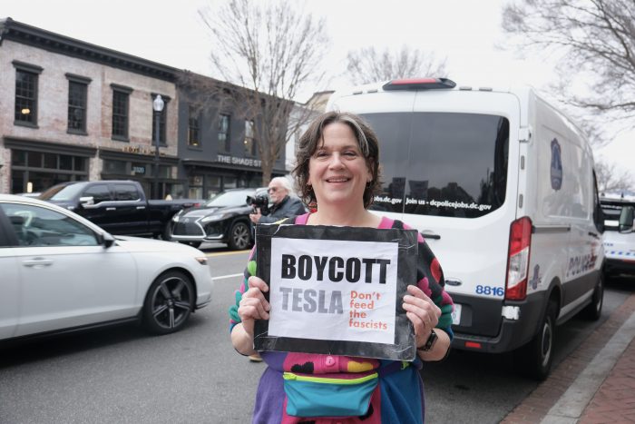 Steffens stands adjacent to the street in a multicolored street holding a paper sign that says, “BOYCOTT TESLA—Don’t feed the facists.”