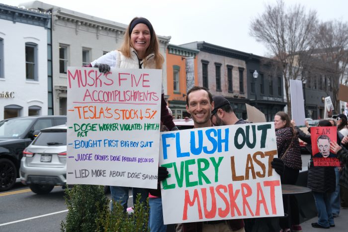 Hollins stands and poses with another demonstrators. Hollins, on the right, holds a large sign that says, “Flush out every last Muskrat.” The woman in a white jacket to his left holds a sign reading, “Musk’s five accomplishments: - TESLA’S STOCK DOWN 5% - DOGE WORKER VIOLATED HATCH -BOUGHT FIRST BIGBOY SUIT - JUDGE ORDERS DOGE PRODUCE RECORDS - LIED MORE ABOUT DOGE SAVINGS.”