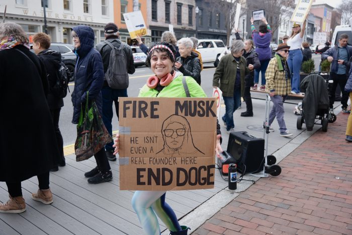 Lowe, wearing yellow neon jacket and an American flag bandana, poses with her cardboard sign that reads, “FIRE MUSK; He isn’t even a real founder here; END DOGE” Next to the text is a drawing of Damien from Mean Girls with a sweatshirt cinched tightly around his face.