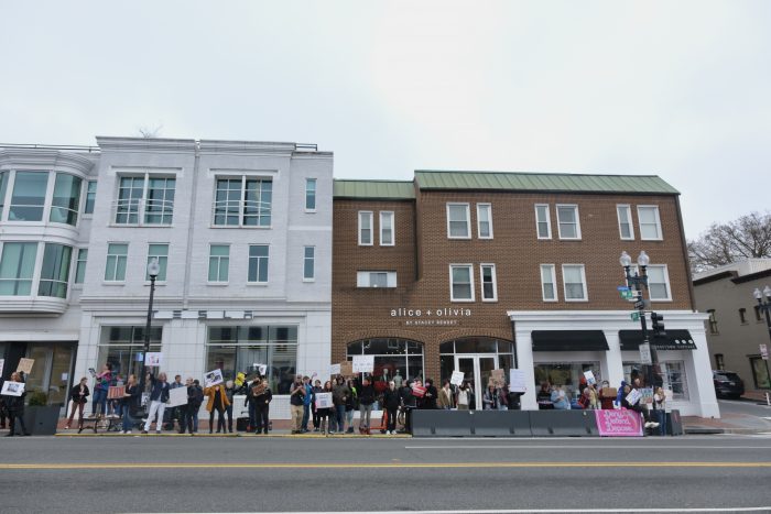 A crowd of around 50 demonstrators gathers holding signs in front of the white Tesla storefront and a brick building with the worlds “Alice + Olivia” on the front.